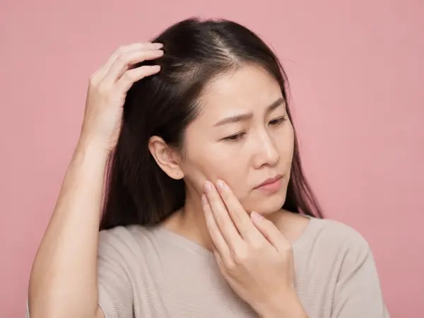 Asian woman with long dark hair touching her cheek while looking thoughtful against a pink background.