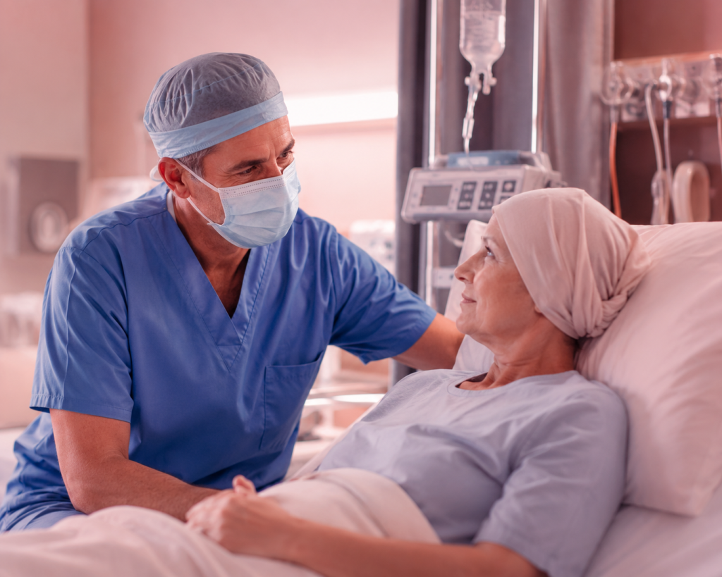 A nurse in blue scrubs and mask sits beside a patient in a hospital bed, offering comfort and support. The patient wears a headscarf and gown and looks up at the caregiver.