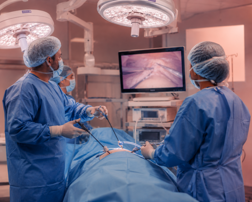 Surgeons in blue scrubs and masks perform laparoscopic surgery with a monitor displaying the internal view of the patient's abdomen.