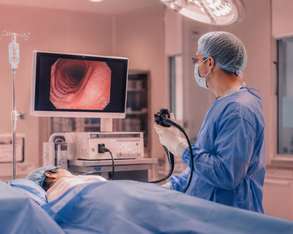 Surgeon in blue scrubs and cap performing an endoscopy; monitor shows the inside of a patient’s airway.