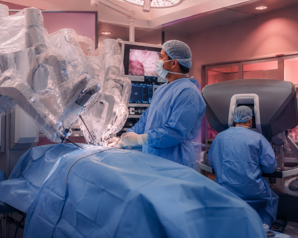 Robot-assisted surgery in a sterile OR with multiple robotic arms around the patient and a monitor displaying images.