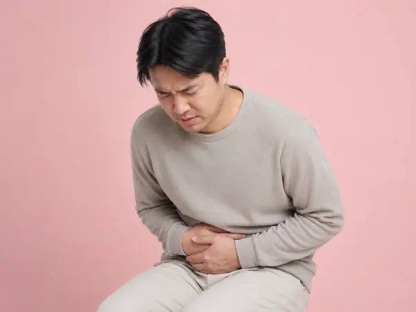 Man sits against a pink background, grimacing in pain while clutching his stomach.