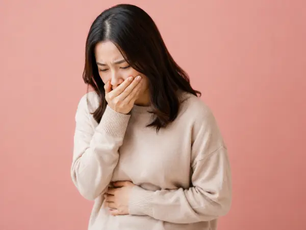 Woman with shoulder-length dark hair in a beige sweater covers her mouth with one hand and holds her stomach, looking unwell against a pink background.