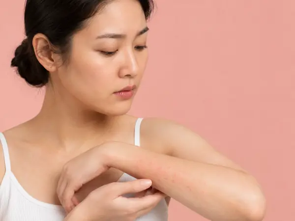 Woman in a white tank top applies lotion to her forearm against a pink background, focused on skincare.