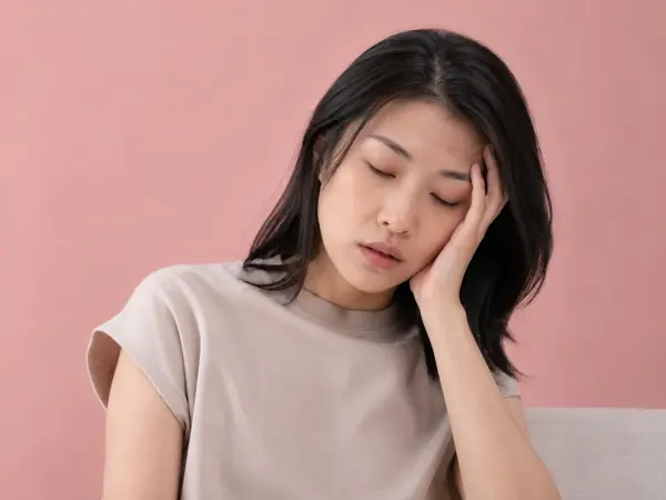 Woman resting her head on her left hand with eyes closed, looking tired against a pink background.